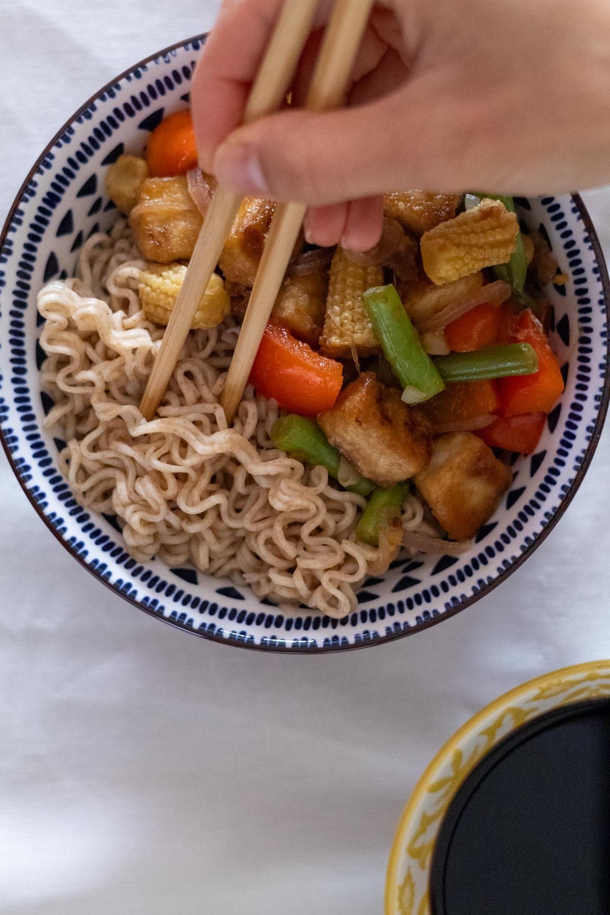 A hand takes noodles with chopsticks from the blue bowl filled with noodles and stir-fried tofu, green beans, red bell pepper, baby corn and red onion.