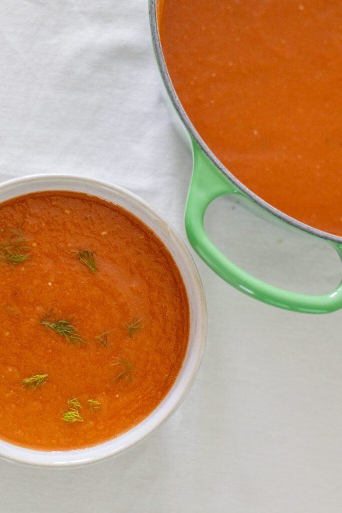 A small white bowl filled with Tomato and Fennel Soup and a large green pot filled with the same soup on the side.