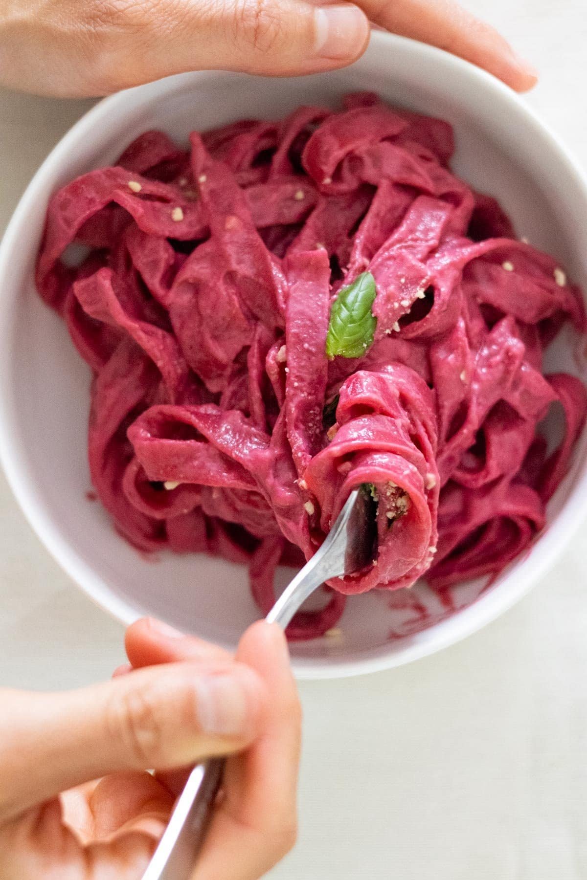 A hand holds a white bowl filled with tagliatelle covered in beetroot sauce, another hand turns pasta with a fork.