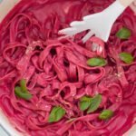 Tagliatelle with a beetroot pasta sauce in a large pot topped with fresh basil and vegan parmesan cheese.