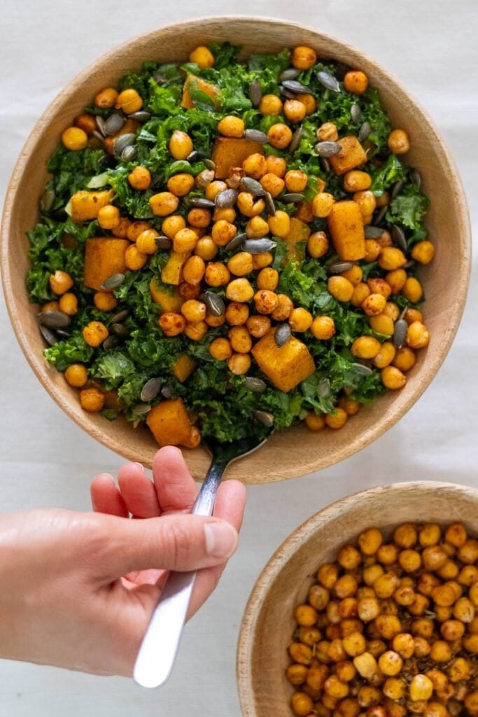 A hand puts a spoon in a wooden bowl filled with Kale, pumpkin seeds and roasted pumpkin and chickpeas.