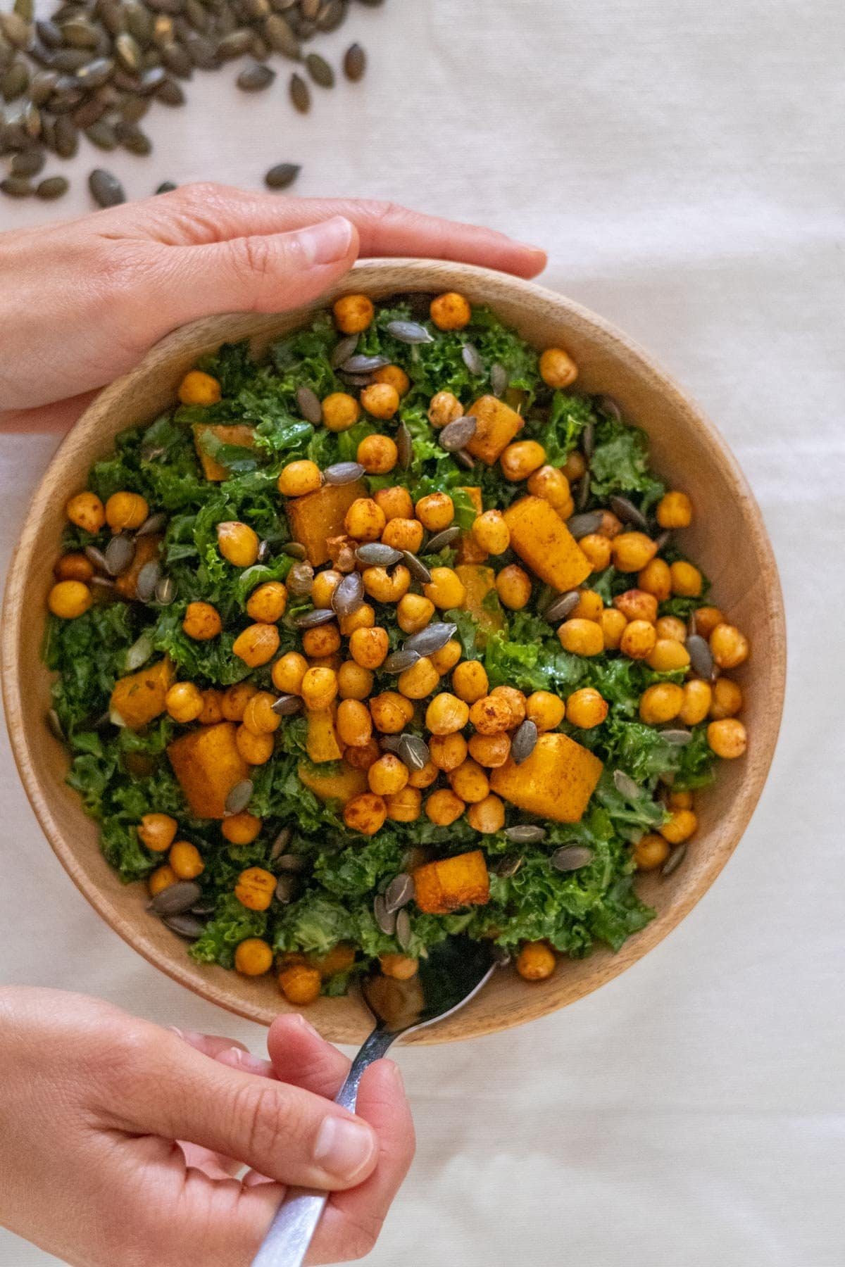 A hand puts a spoon in a wooden bowl filled with Kale, pumpkin seeds and roasted pumpkin and chickpeas.