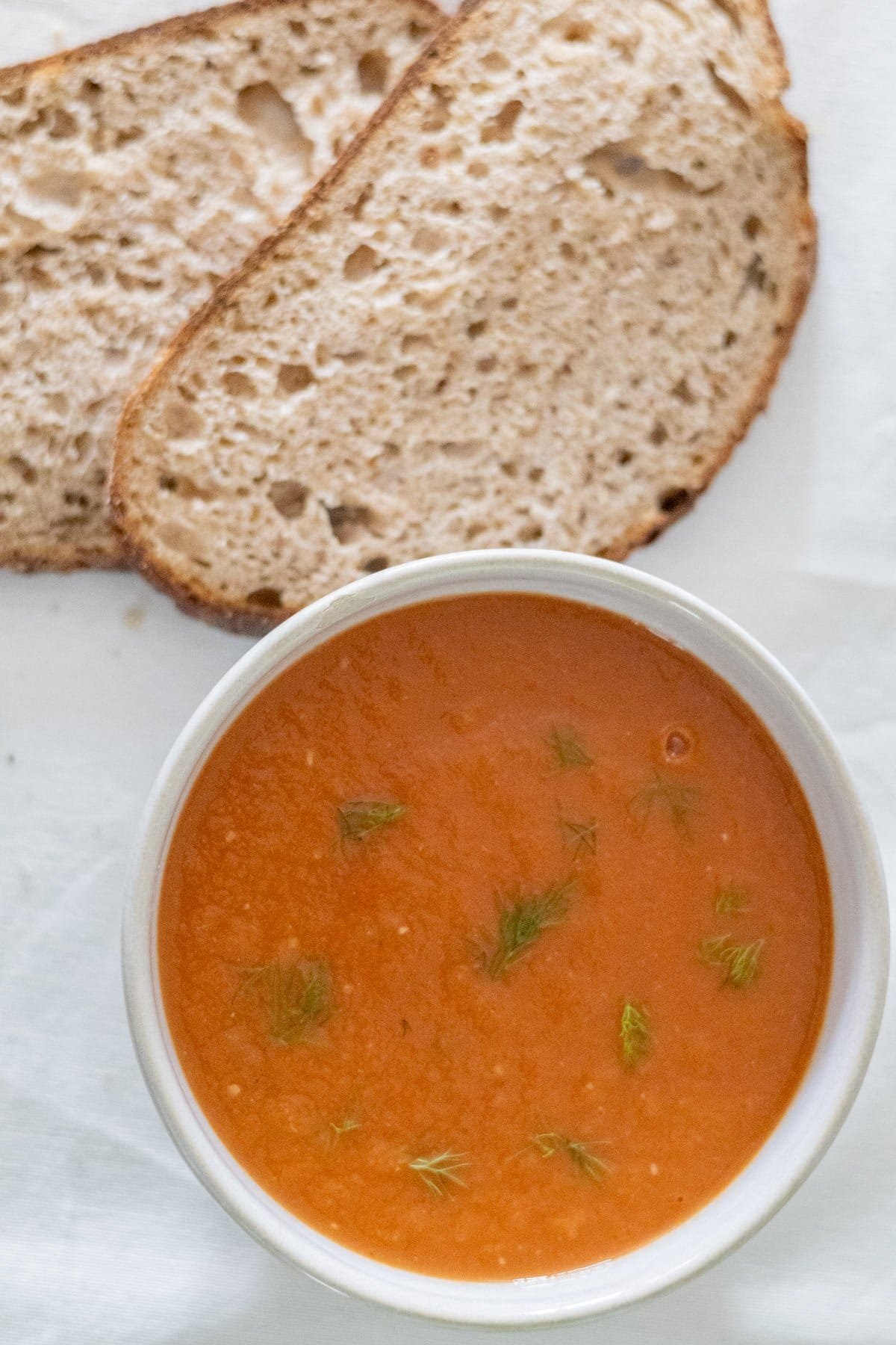 A small white bowl filled with Tomato and Fennel Soup with two slices of sourdough bread on the side.