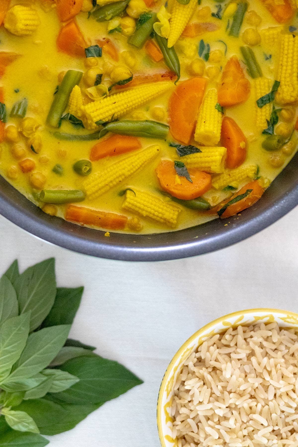 Vegan Thai Yellow Curry with green beans, carrot, baby corn and chickpeas served in a black wok with cooked rice and fresh Thai basil on the side.