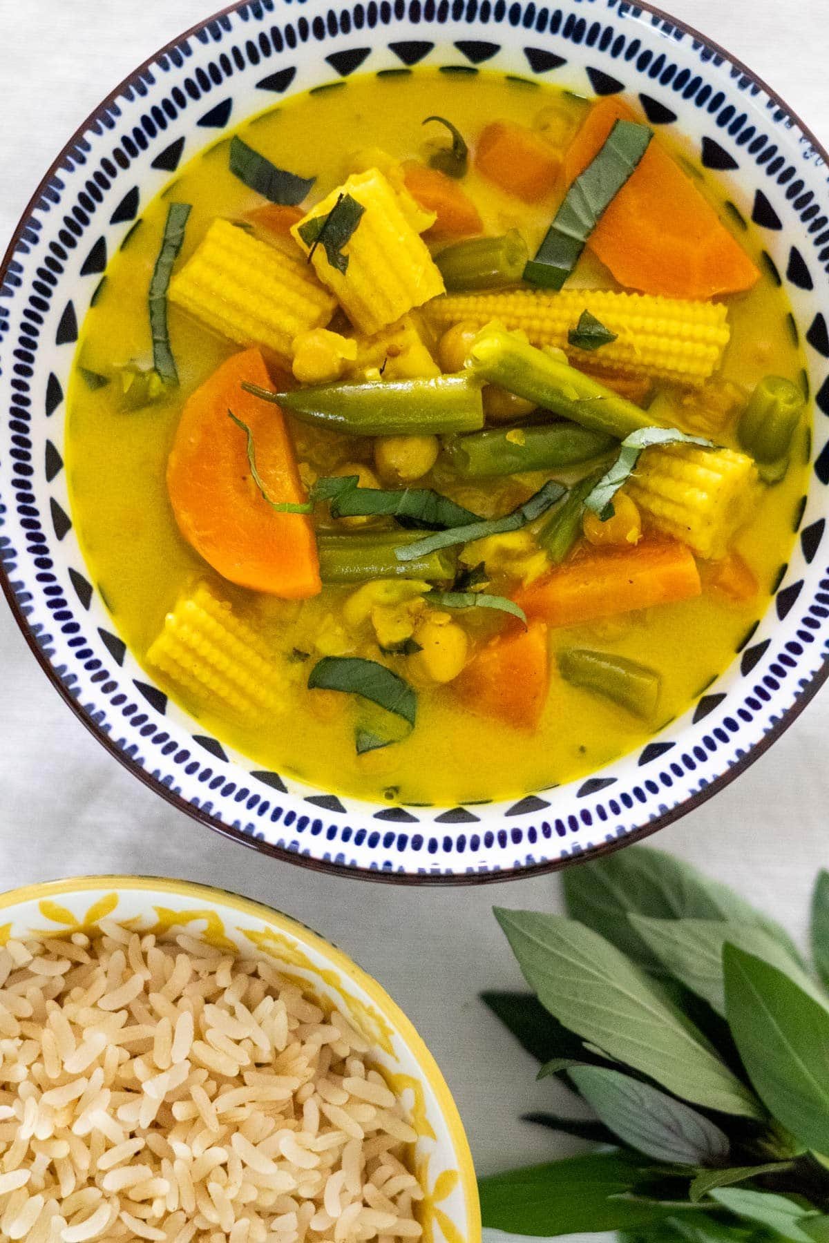 Vegan Thai Yellow Curry with green beans, carrot, baby corn and chickpeas served in a blue and white bowl with cooked rice and fresh Thai basil on the side.