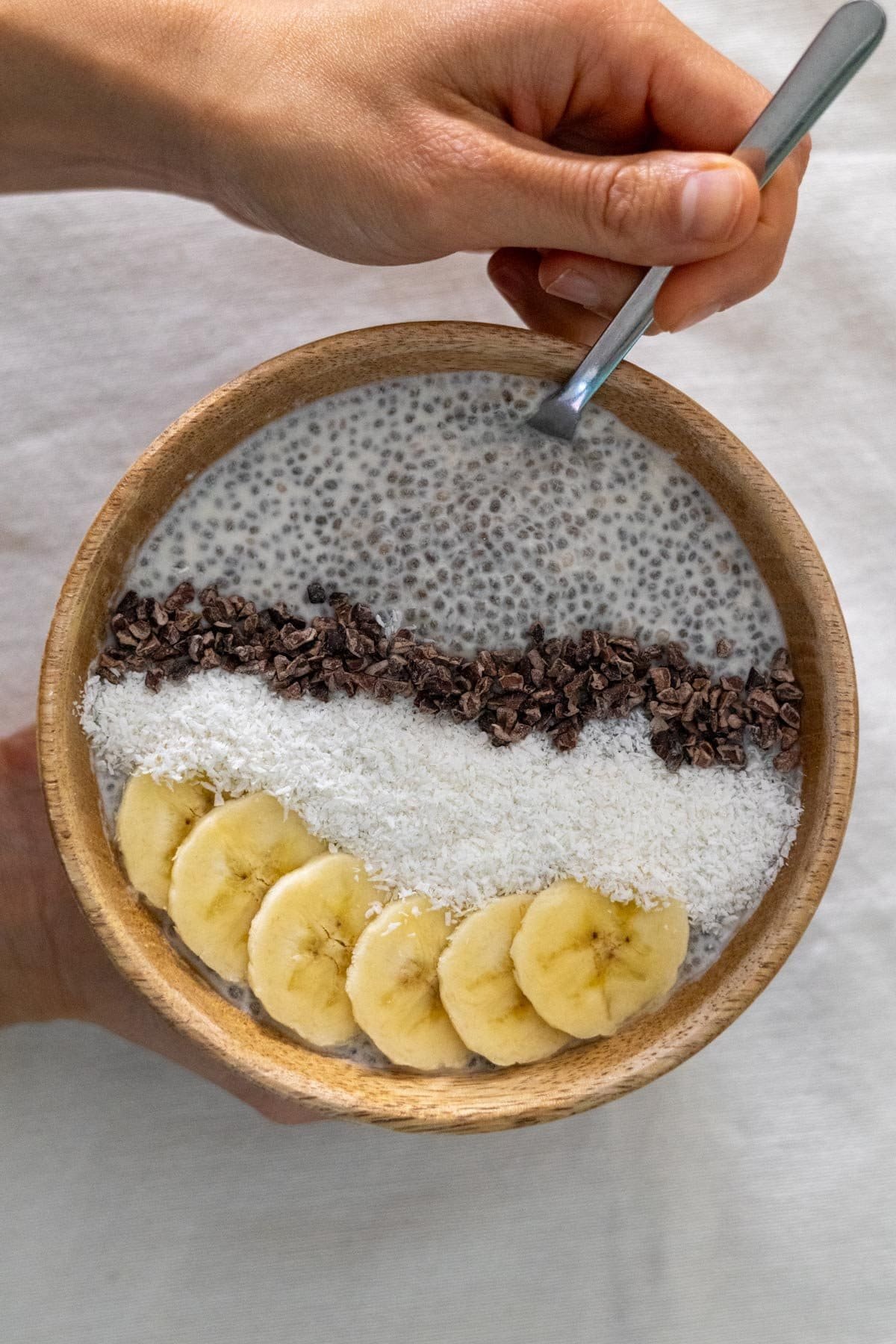 Coconut Chia Pudding in a wooden bowl topped with cocoa nibs, grated coconut and banana.