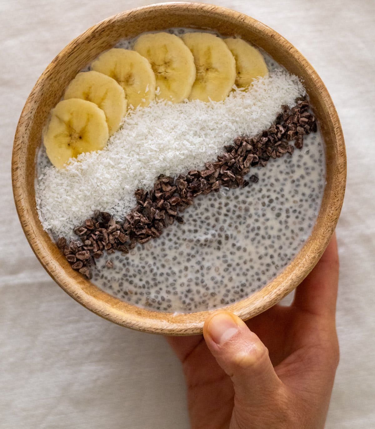 A hand holds a wooden bowl filled with coconut chia pudding topped with cocoa nibs, grated coconut and banana.