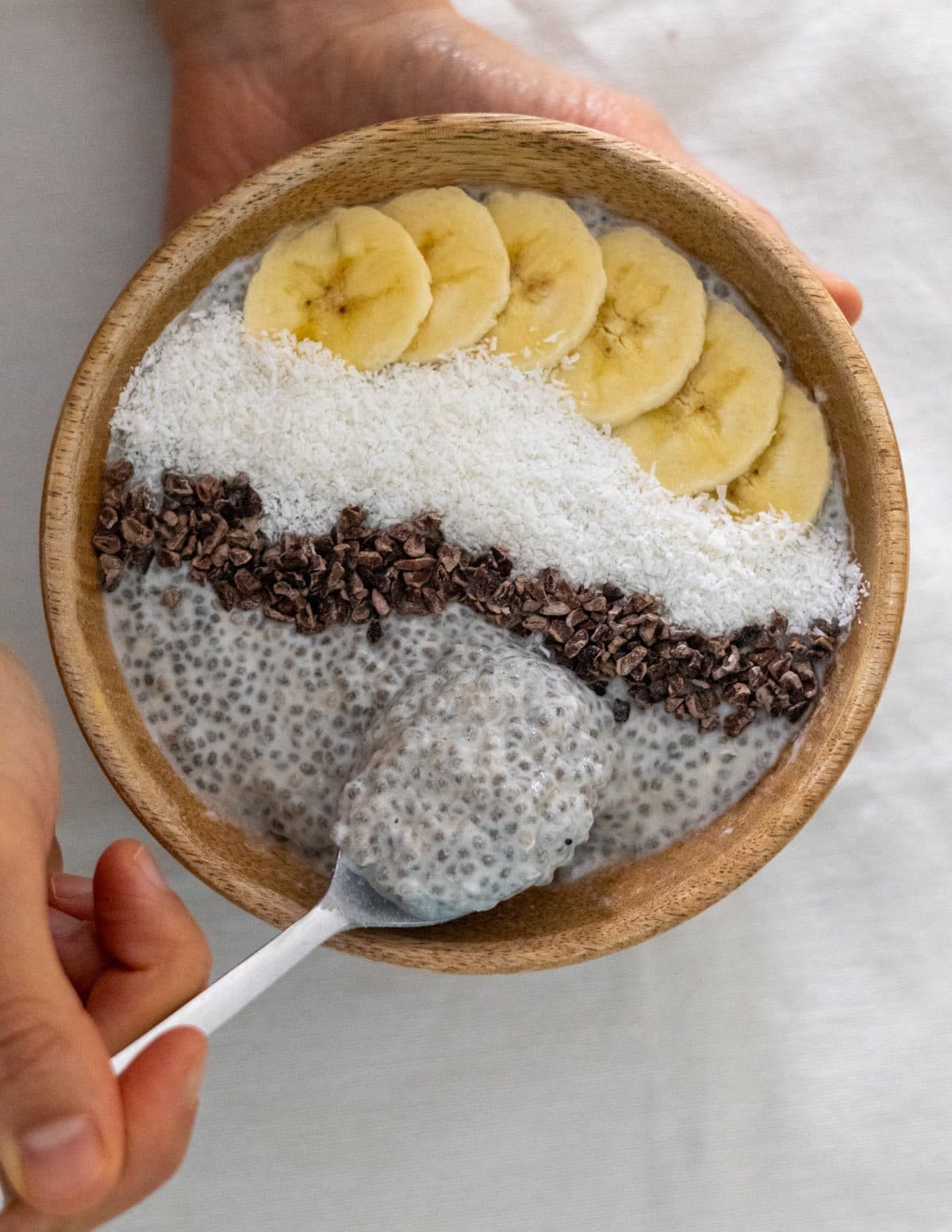 A hand holds a spoon in a wooden bowl filled with coconut chia pudding.