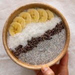 Creamy coconut chia pudding in a wooden bowl topped with banana, cocoa nibs and grated coconut.