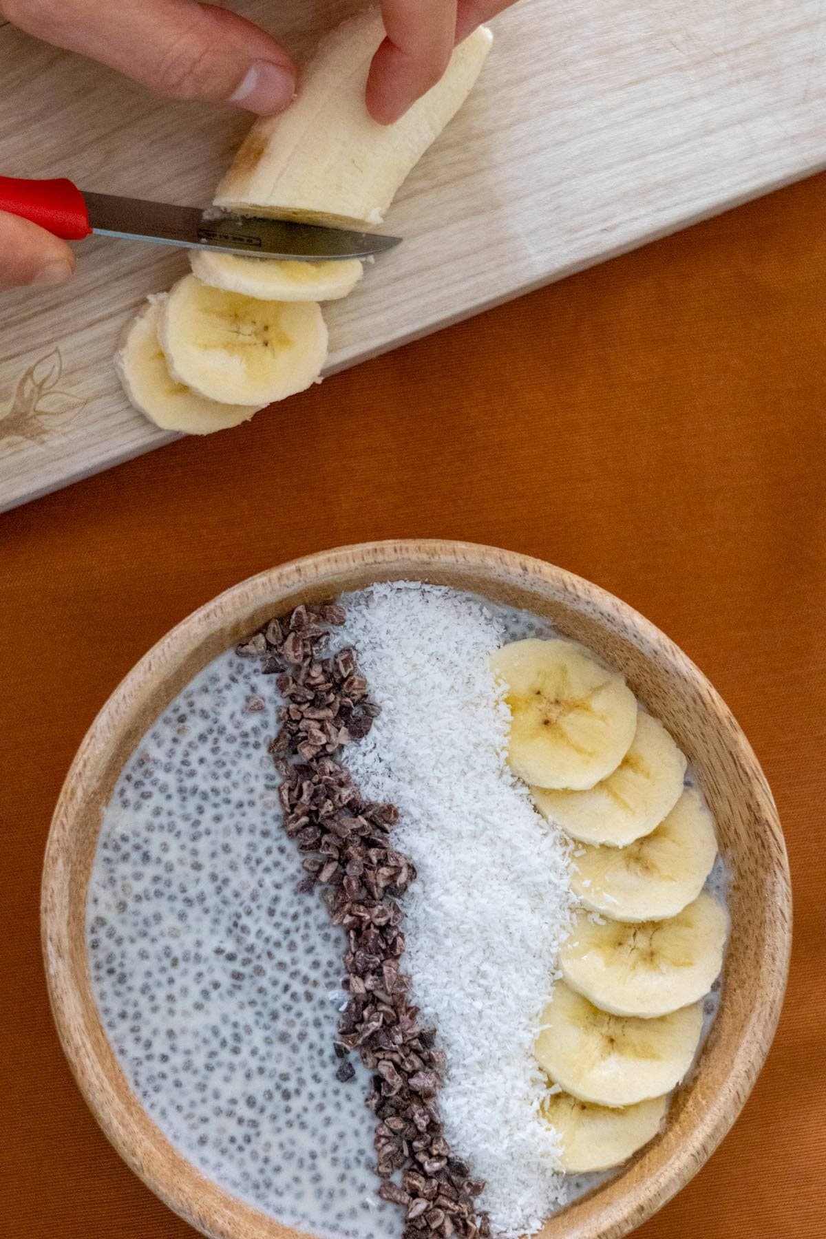 Creamy coconut chia pudding in a wooden bowl with a hand cutting a banana on a wooden cutting board on the side.