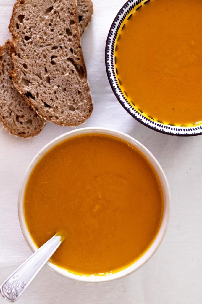 Two bowls filled with carrot and lentil soup with a spoon in one of the bowl. Served with sourdough bread on the side.