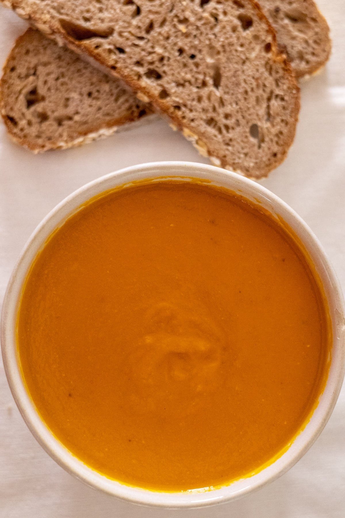 A white bowl filled with carrot and lentil soup with some bread on the side.