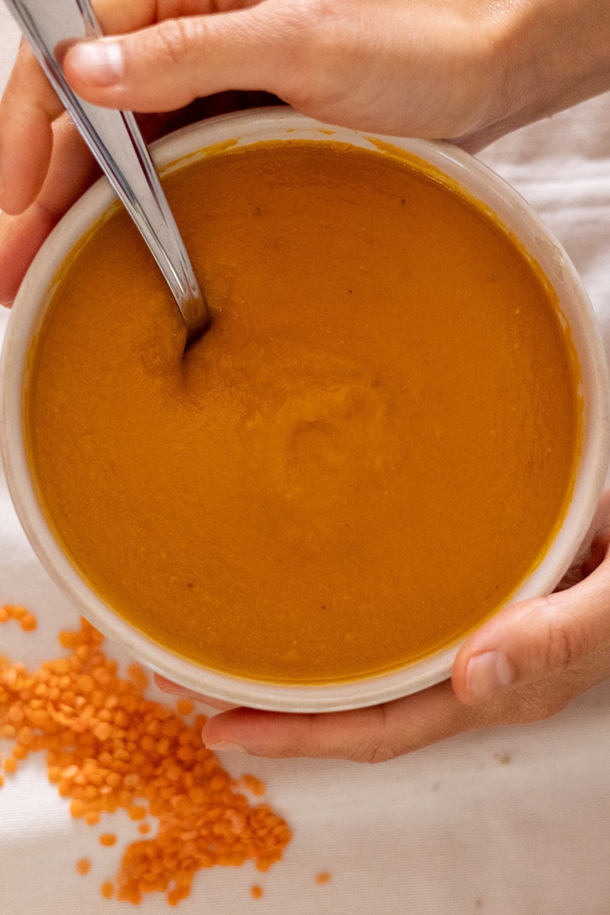 Two hands hold a white bowl filled with carrot and lentil soup.