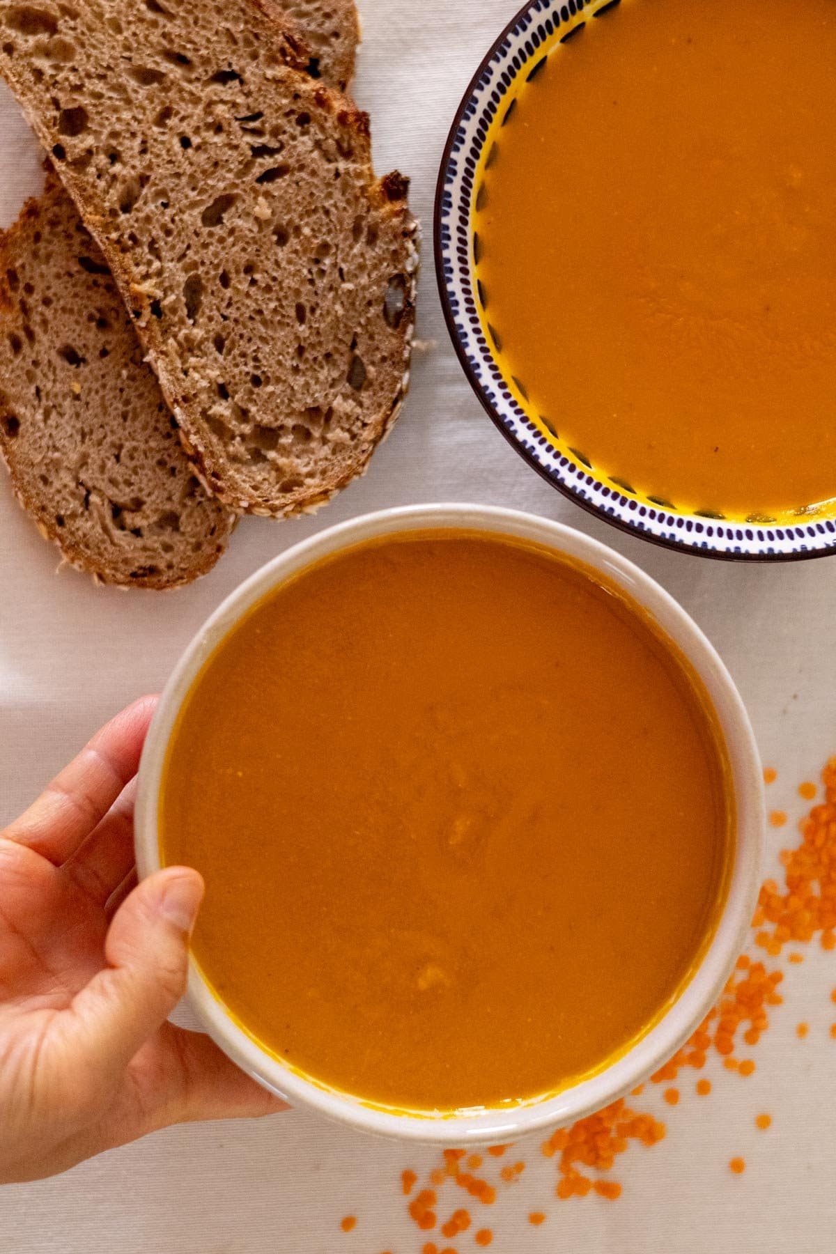 Two bowl filled with carrot and lentil soup with some bread and dried red lentils on the side.