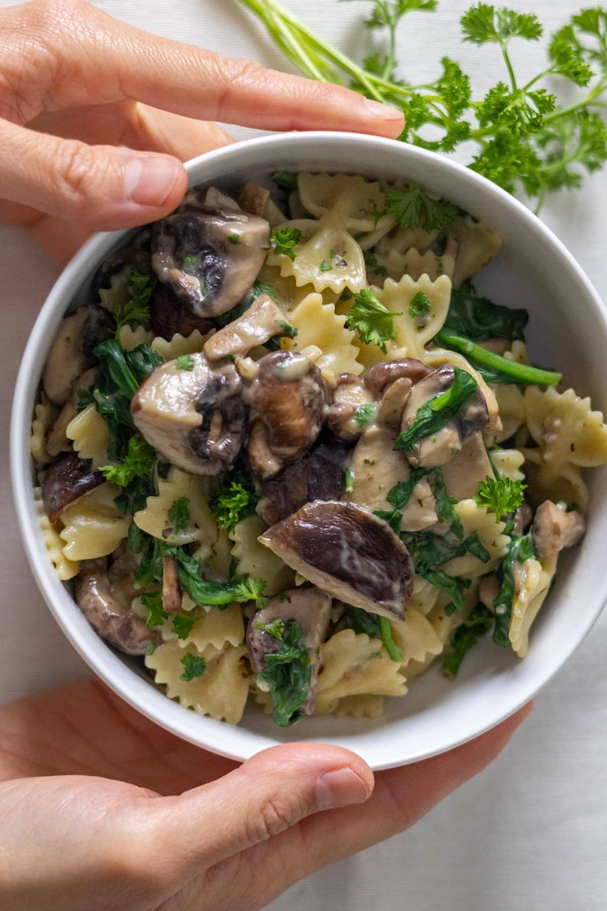 Two hands hold a white bowl filled with cooked mushrooms, spinach, parsley and pasta.