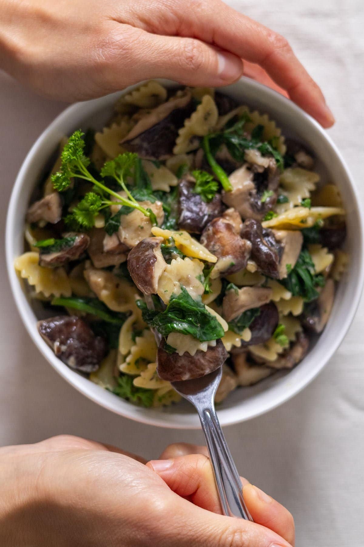 One hand holds a white bowl filled with Spinach and Mushroom Pasta, the other hand takes some of the Spinach and Mushroom Pasta with a fork.