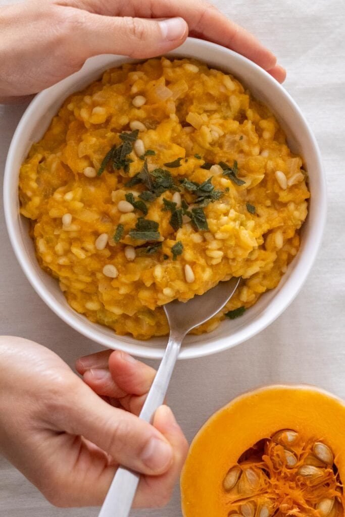 A hand holds a small white bowl filled with Vegan Pumpkin and Sage Risotto, another hand takes risotto from the bowl with a spoon.