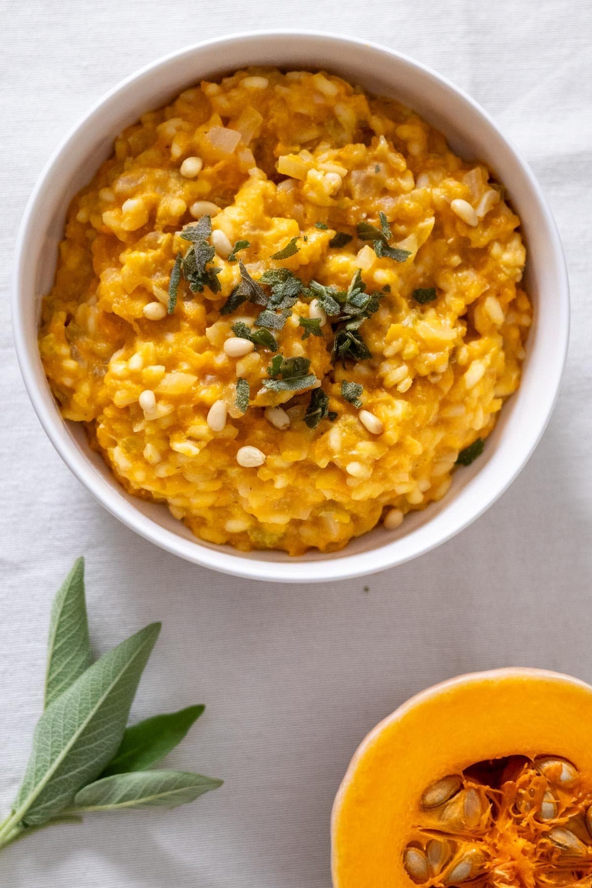 Pumpkin and Sage Risotto in a small white bowl topped with fried sage and pine nuts with some fresh sage and half a pumpkin on the side.