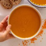 a hand holds a white bowl filled with carrot and lentil soup.