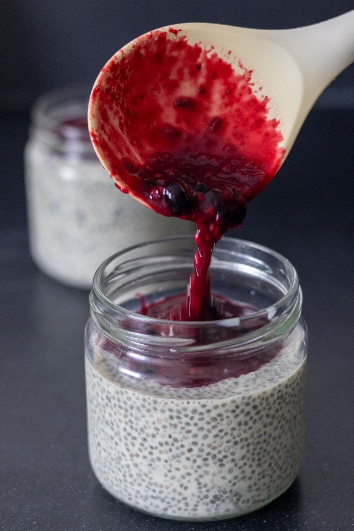 Warm berry sauce is poured into a small mason jar filled with chia pudding using a white serving spoon.