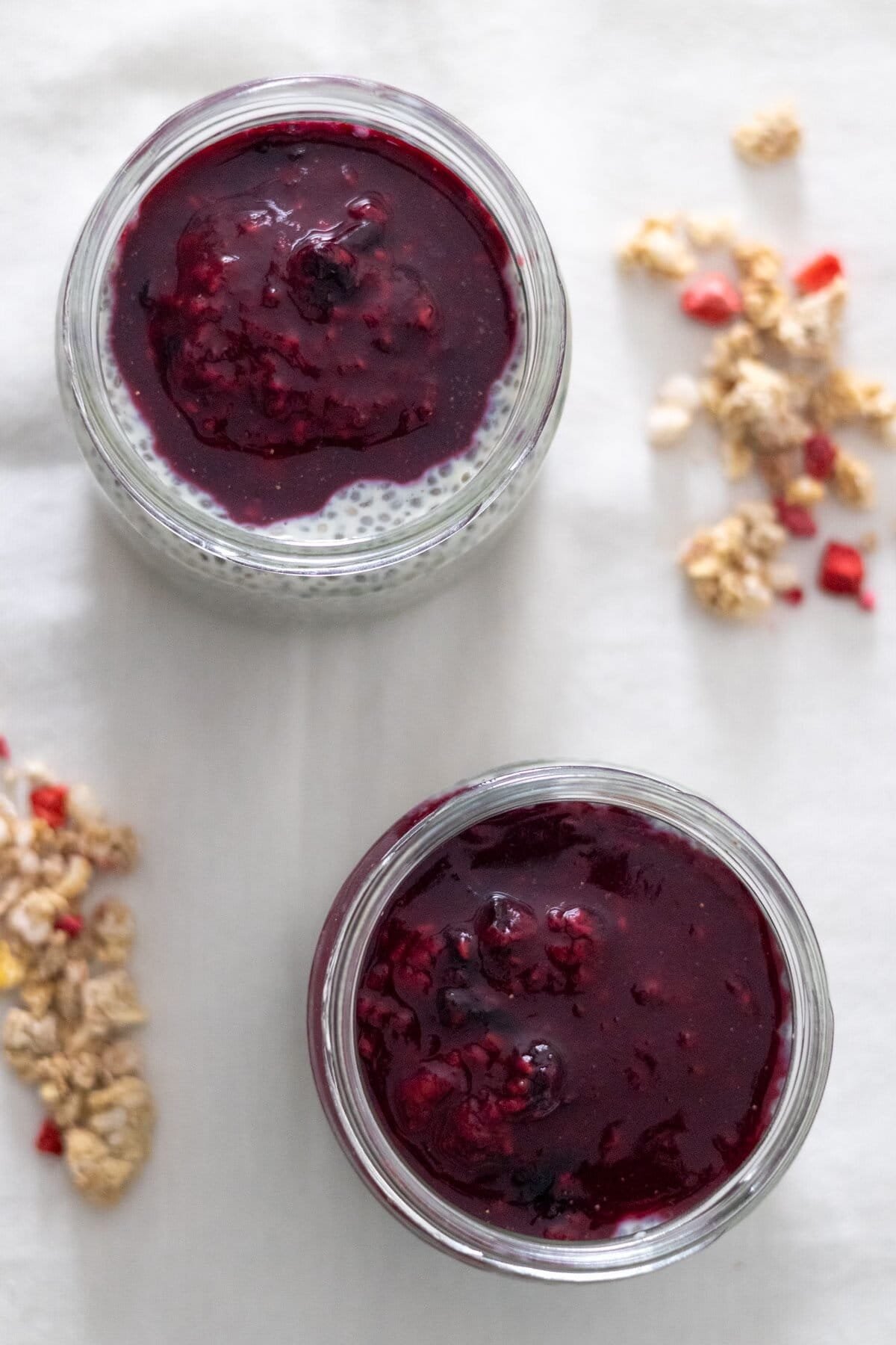 Two small mason jars filled with chia pudding topped with a sauce made of raspberries and blueberries with some granola on the side.