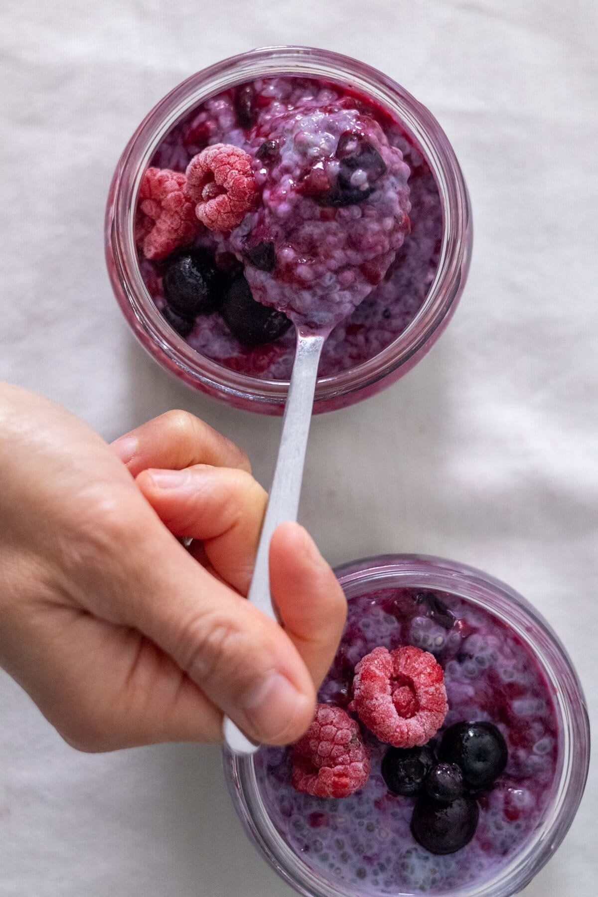 Two small mason jars filled with Berry Chia Pudding. A hand takes a spoonful of berry chia pudding from one jar.