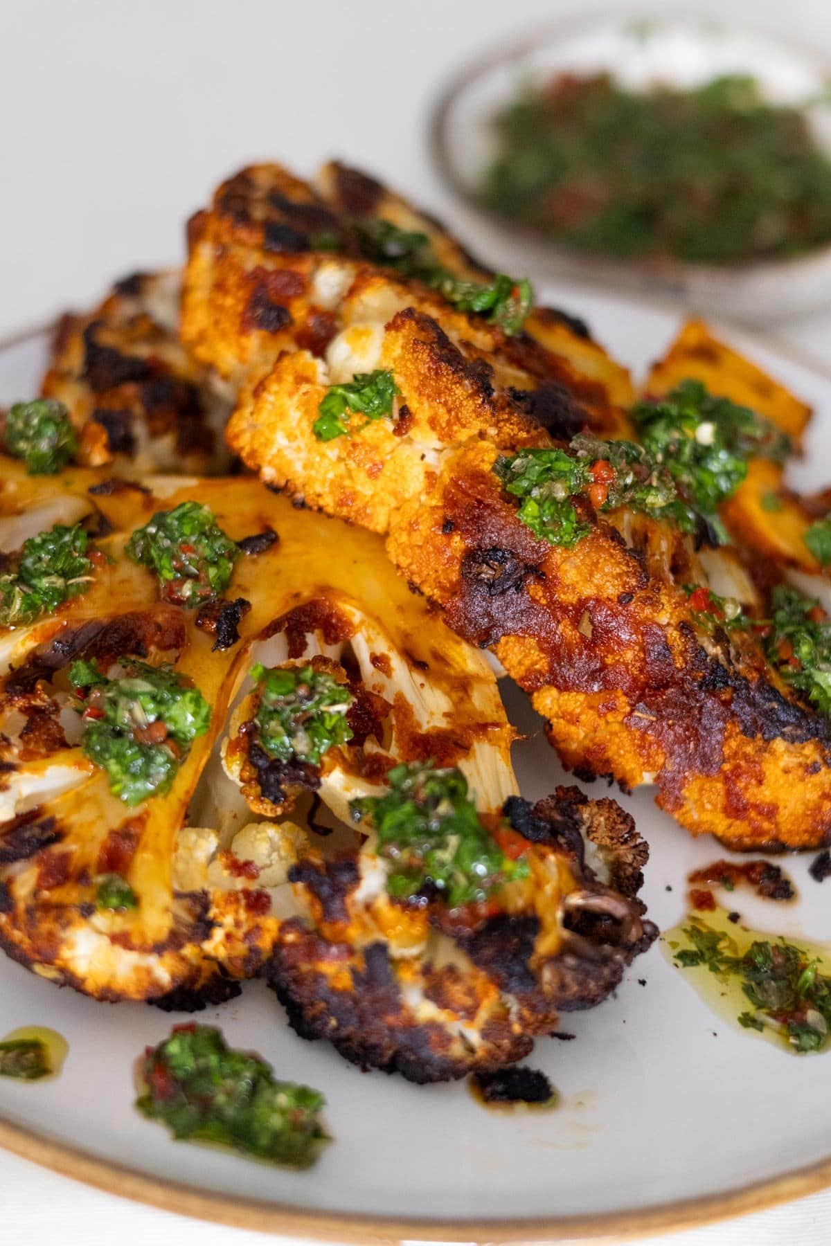 Two Vegan Cauliflower Steaks on a white plate topped with Chimichurri with more Chimicurri in the back.