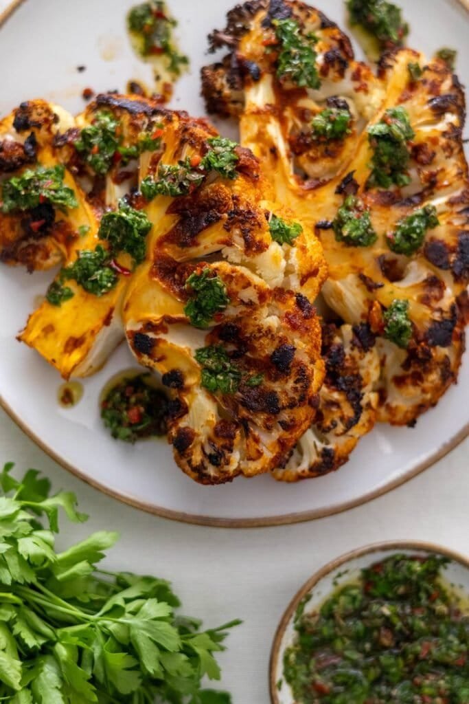 Two Vegan Cauliflower Steaks on a white plate topped with Chimichurri with more chimichurri and fresh parsley on the side.